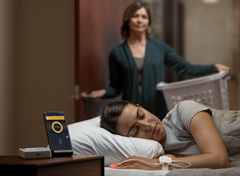 Young sleeping person monitored by Masimo Opioid with older person behind holding a laundry basket. 